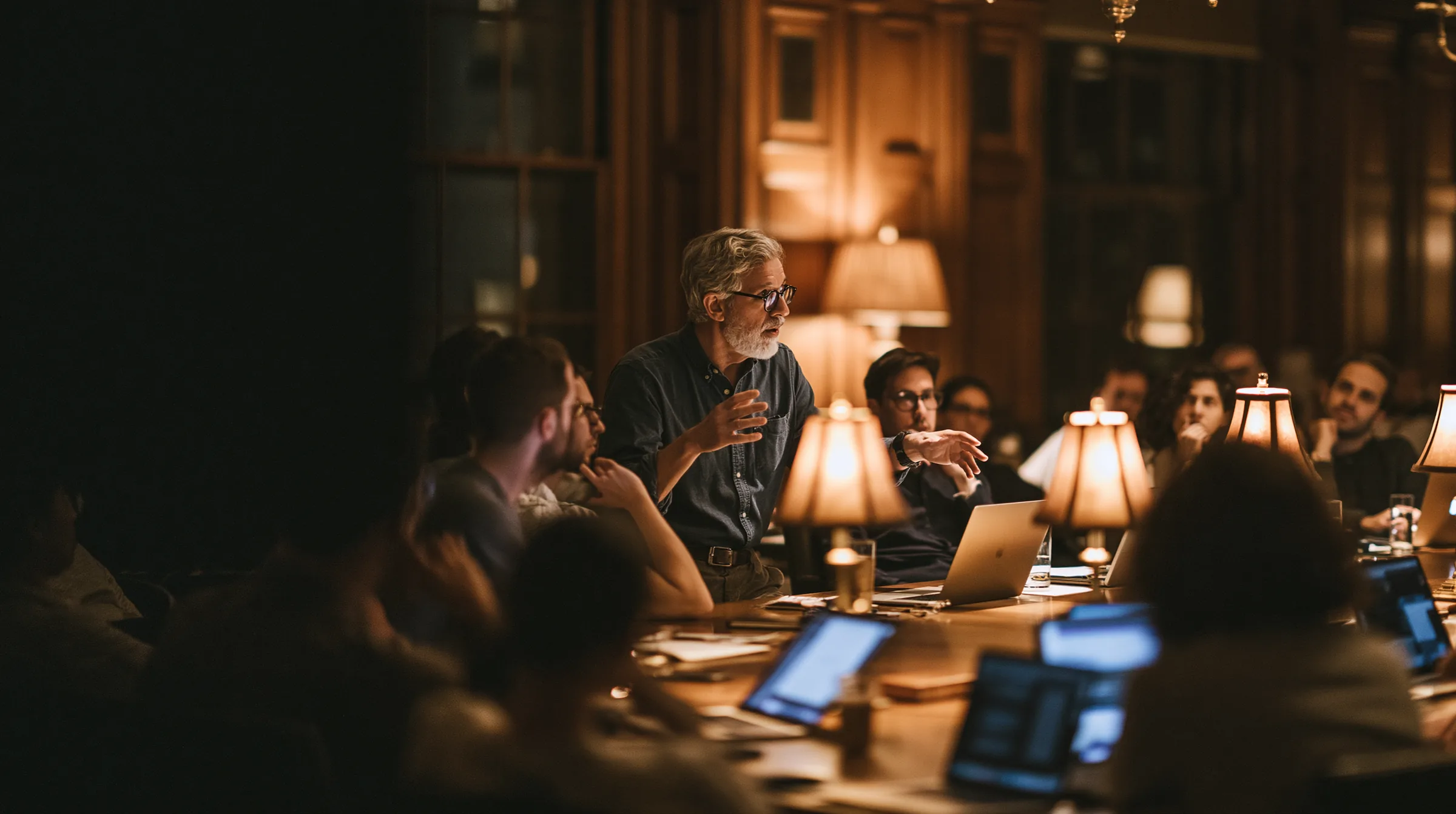 An evening university seminar in a wood-paneled room — adults of varied ages around a long table with laptops, lit by table lamps
